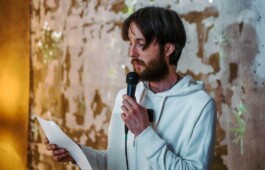 Man in a white hoodie reads from a paper while holding a microphone. The background wall is textured and rustic.