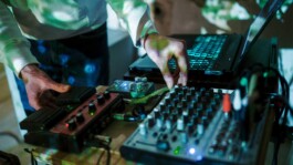 A person operates a sound mixing board and effects pedals on a table, illuminated by colorful projected light patterns.