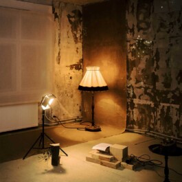 Performance installation. A warmly lit room with peeling wallpaper, a glowing floor lamp, scattered papers and stacked wooden blocks.